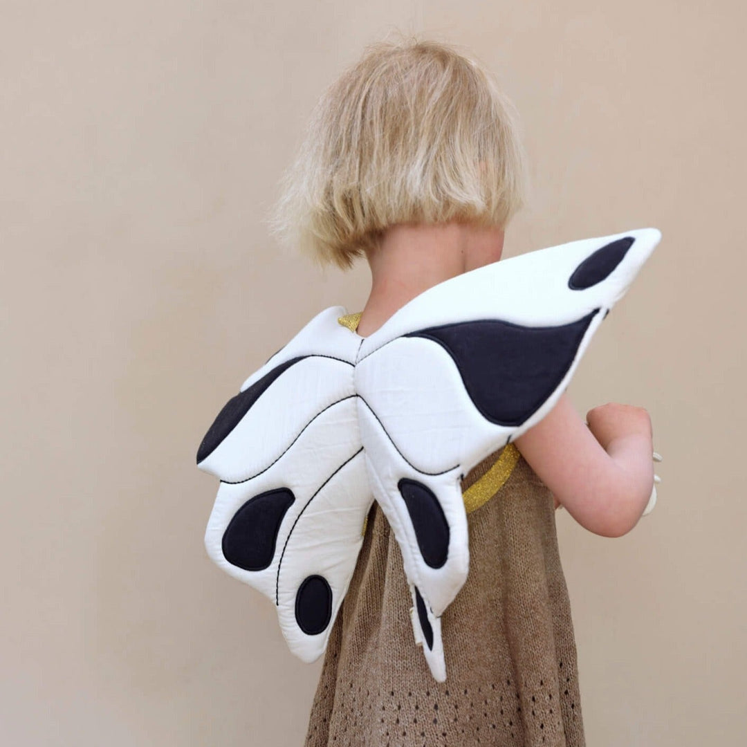 Girl with short blonde hair wearing a pair of white and black butterfly wings. She has her back to the camera. 